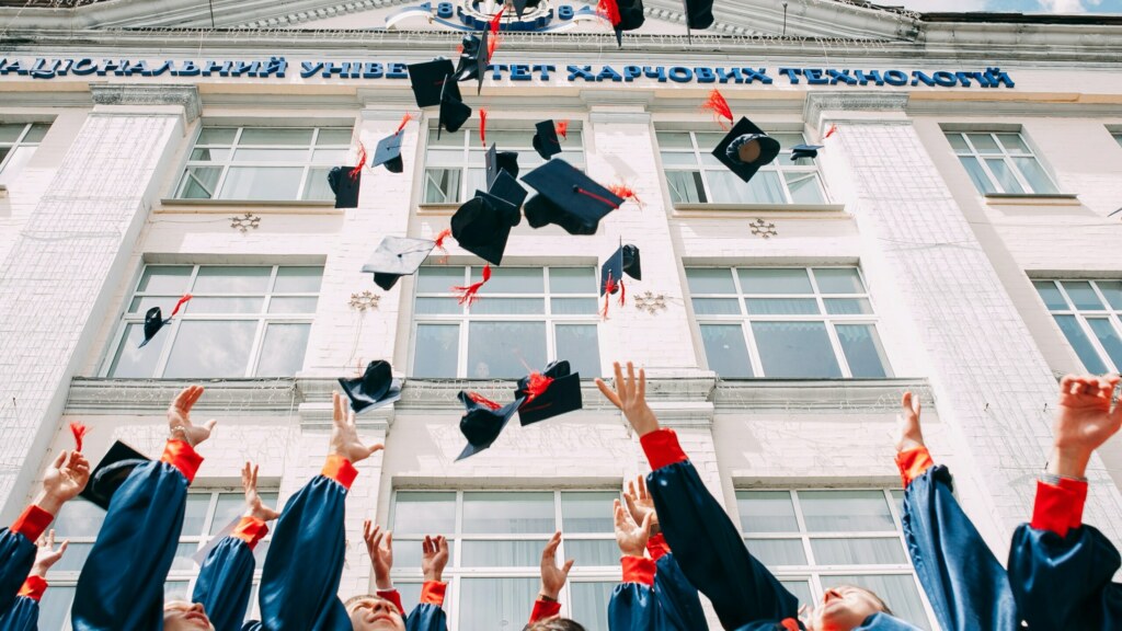 Photo of young people in their graduating attire, throwing their hats in the air in celebration outside their University building
