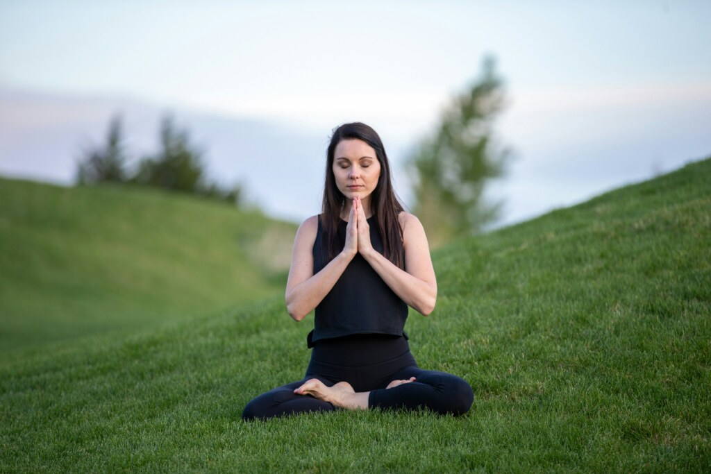Picture of a young women dressed in black practicing meditation on grass with her two hands joined up in front of her chest and her legs crossed.