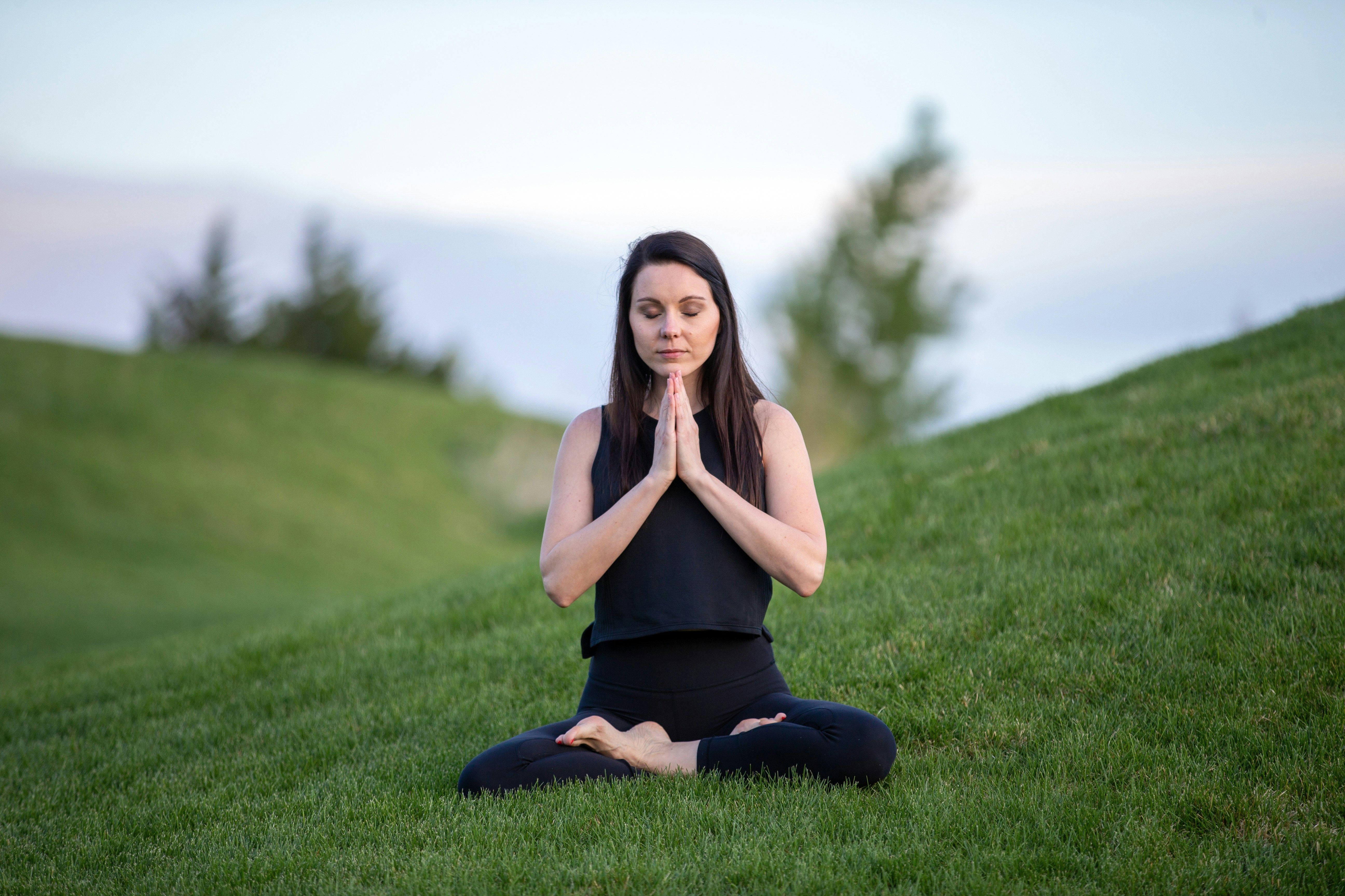 Picture of a young women dressed in black practicing meditation on grass with her two hands joined up in front of her chest and her legs crossed.